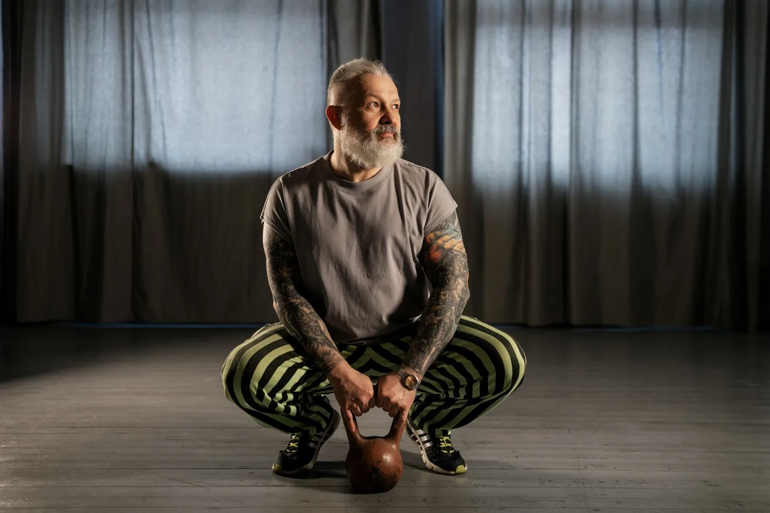 Older bearded man holding a kettlebell in a deep squat in a dim gym